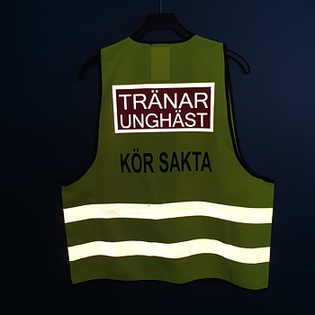 Reflexväst färg gul, Text bak på västen TRÄNAR UNGHÄST i ljusgrå reflex i en rödplatta med reflexram runt. svart text under KÖR SAKTA