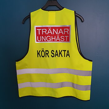 Reflexväst färg gul, Text bak på västen TRÄNAR UNGHÄST i ljusgrå reflex i en rödplatta med reflexram runt. svart text under KÖR SAKTA text fram UNGHÄST RÄDD .