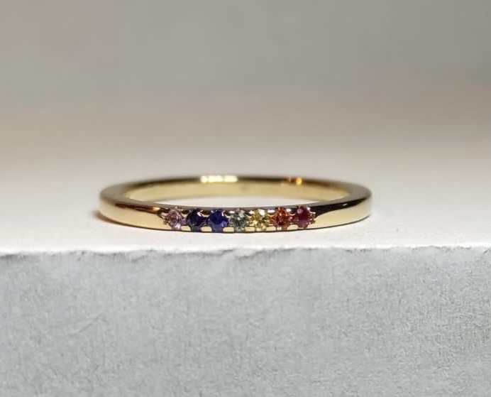 Ring Ellinor i 18K guld med rubin och safirer i regnbågsfärger, liggande mot ljus bakgrund