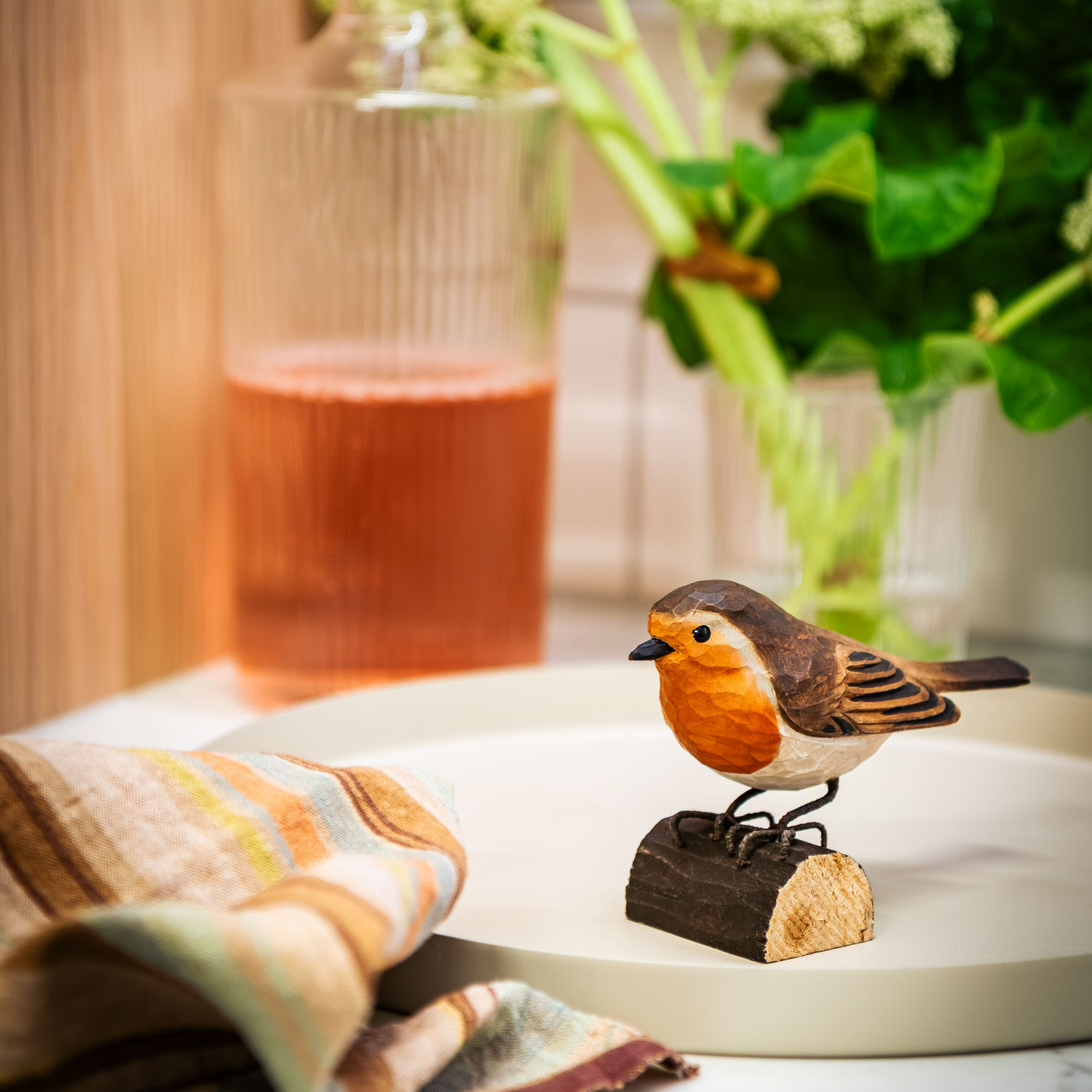 DecoBird Rouge-gorge posé sur un plateau près d’une serviette rayée, d’une carafe en verre avec une boisson et de plantes vertes.