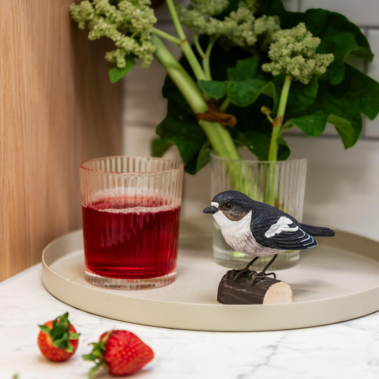 DecoBird Svartvit flugsnappare står på ett fat bredvid ett glas med röd dryck, jordgubbar och gröna växter.