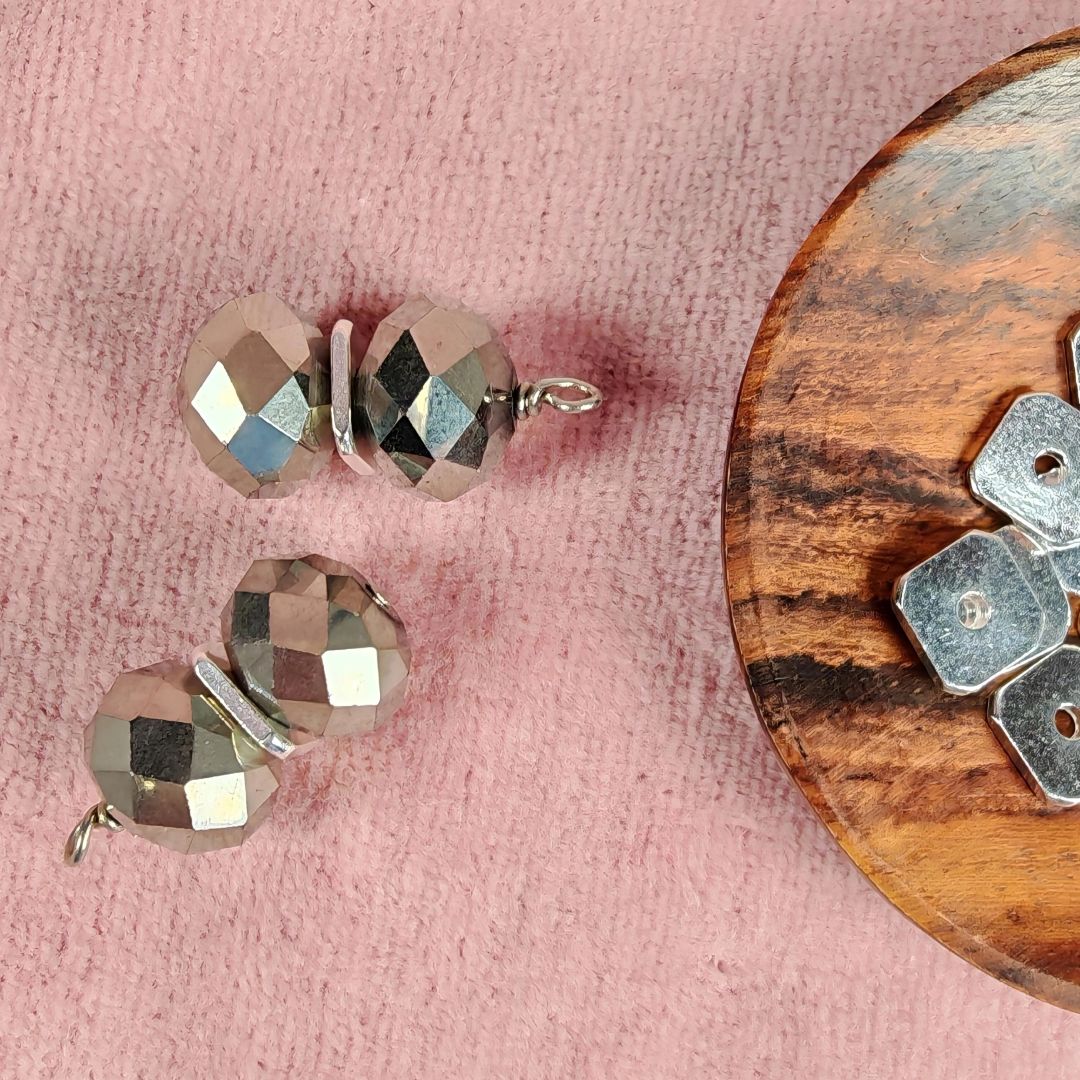 Fyrkantiga silverfärgade mellandelar 8x8 mm med hål på ca 1,5 mm, visade tillsammans med facettslipade pärlor på rosa tyg och en träbricka.