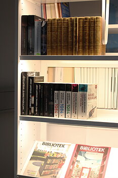 Maggie Hyllebelysning 580 – smal LED-lysskinne montert under hylleplan som gir jevn belysning av bøker; magnetfeste for enkel montering.