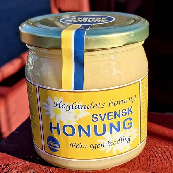 Närbild på en 500 grams glasburk med svensk honung, gulvit etikett med präglat bivaxmönster i glaset och blågul sigillförslutning.