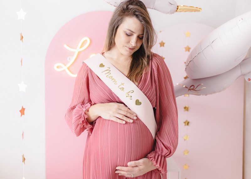 Gravid kvinna bär ljusrosa Mom To Be sash med guldtext under babyshower.