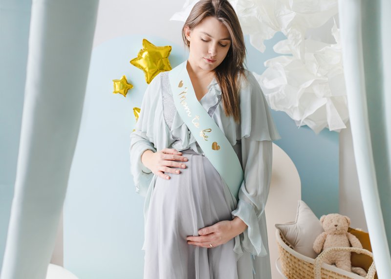 Gravid kvinna bär ljusblå Mom To Be sash med guldtext under babyshower.