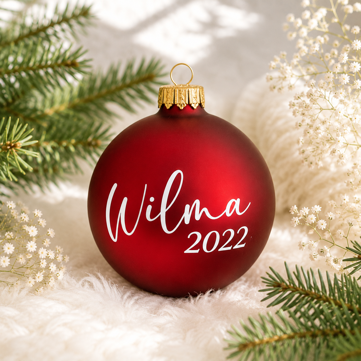 Personlig röd matt julkula i glas med namnet Wilma och årtalet 2022 i vit vinyl, fotograferad i en ljus och stämningsfull julmiljö med granris.