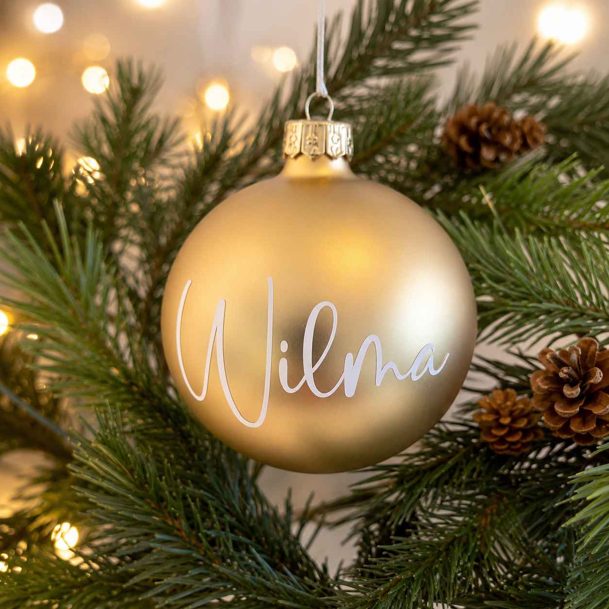 Personlig champagnefärgad matt julkula i glas med namnet Wilma i vit vinyl, fotograferad i en varm och stämningsfull julmiljö med granris, kottar och mjuk julbelysning.