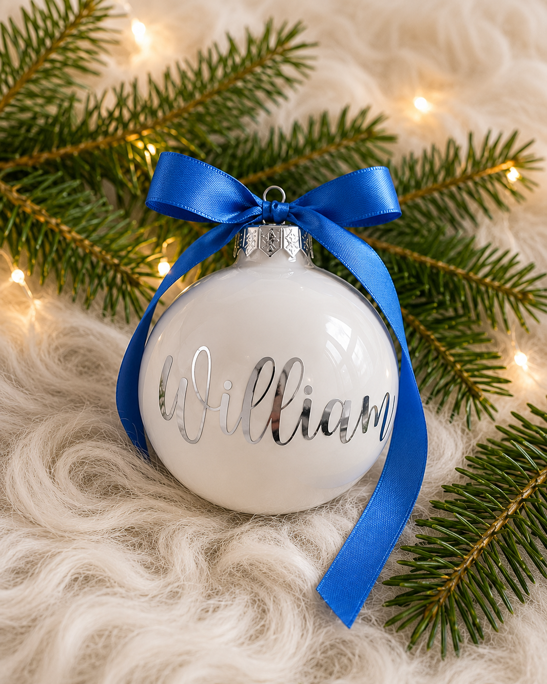 Personlig vit julkula i glas med namnet William i silvertext och blått satinband, omgiven av granris och varm julbelysning
