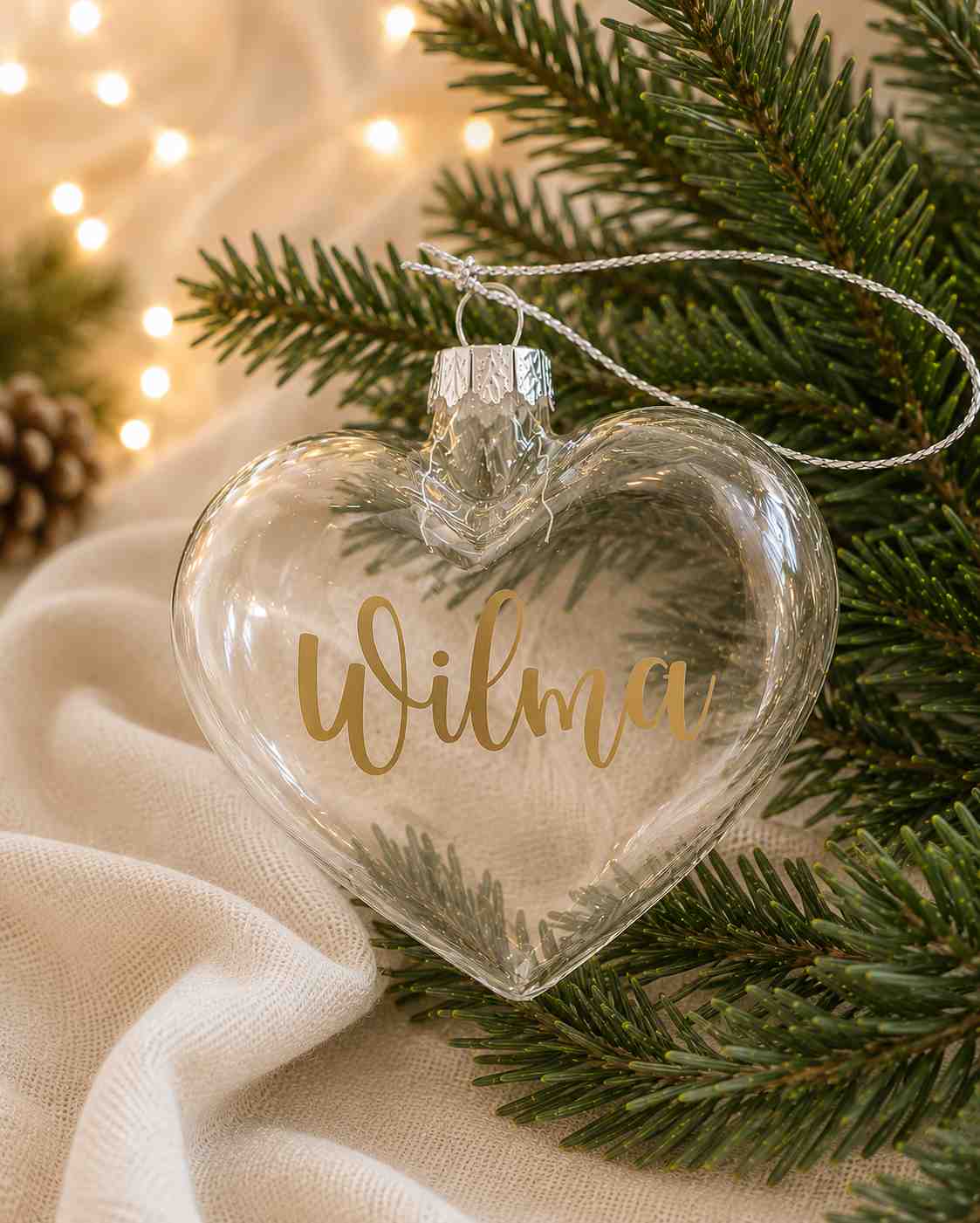 Personlig hjärtformad julkula i transparent munblåst glas med namnet Wilma i guldvinyl, fotograferad i en varm och stämningsfull julmiljö med granris och mjuk julbelysning.