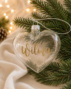 Personlig hjärtformad julkula i transparent munblåst glas med namnet Wilma i guldvinyl, fotograferad i en varm och stämningsfull julmiljö med granris och mjuk julbelysning.
