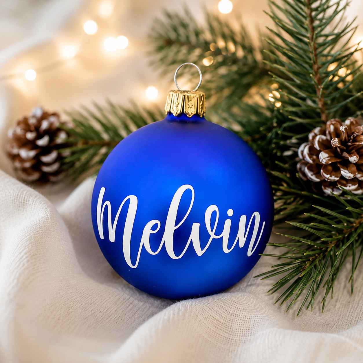 Personlig royalblå matt julkula i glas med namnet Melvin i vit vinyl, fotograferad i en varm och elegant julmiljö med granris, kottar och mjuk belysning.