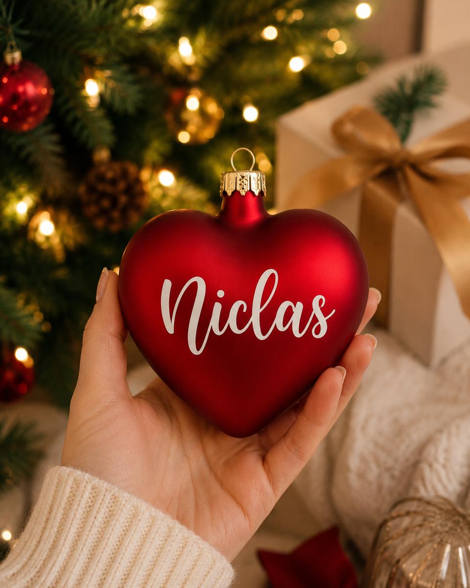 Personlig röd hjärtformad julkula med namn i munblåst glas. Julkulan hålls i handen framför en julgran med varm julbelysning och presenter.