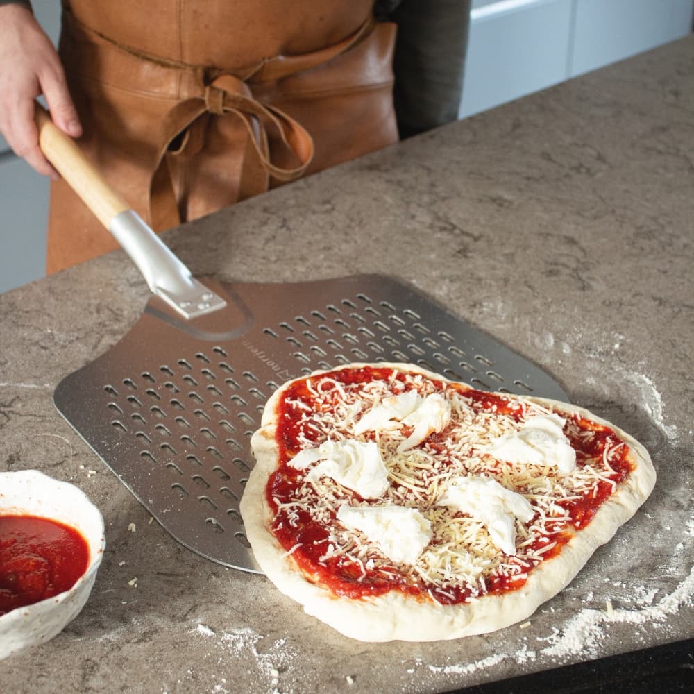 Pizzaspade bruges på ubrændt pizza med tomatsauce og ost af en person med brunt forklæde