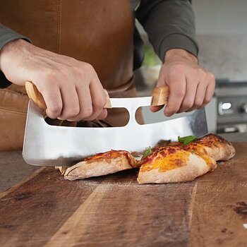 Person med forklæde skærer pizza på skærebræt med pizzaskærer fra norrjern