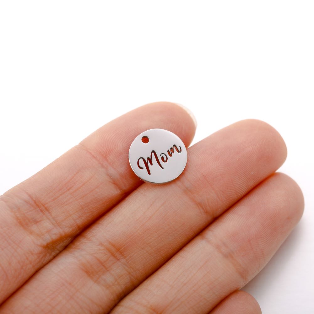 Rund berlock Mom i guldfärgat rostfritt stål, berlock med utskuren text “Mom”, diameter ca 12 mm, visad i närbild.