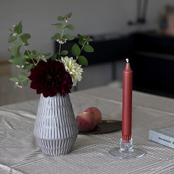 Ett mörkrött kronljus från Liljeholmens står i en låg, transparent ljusstake på ett randigt dukat matbord. Bredvid står en grå keramikvas med mörkröda och ljusa dahlior samt gröna kvistar. I bakgrunden ligger två persikor på ett fat.