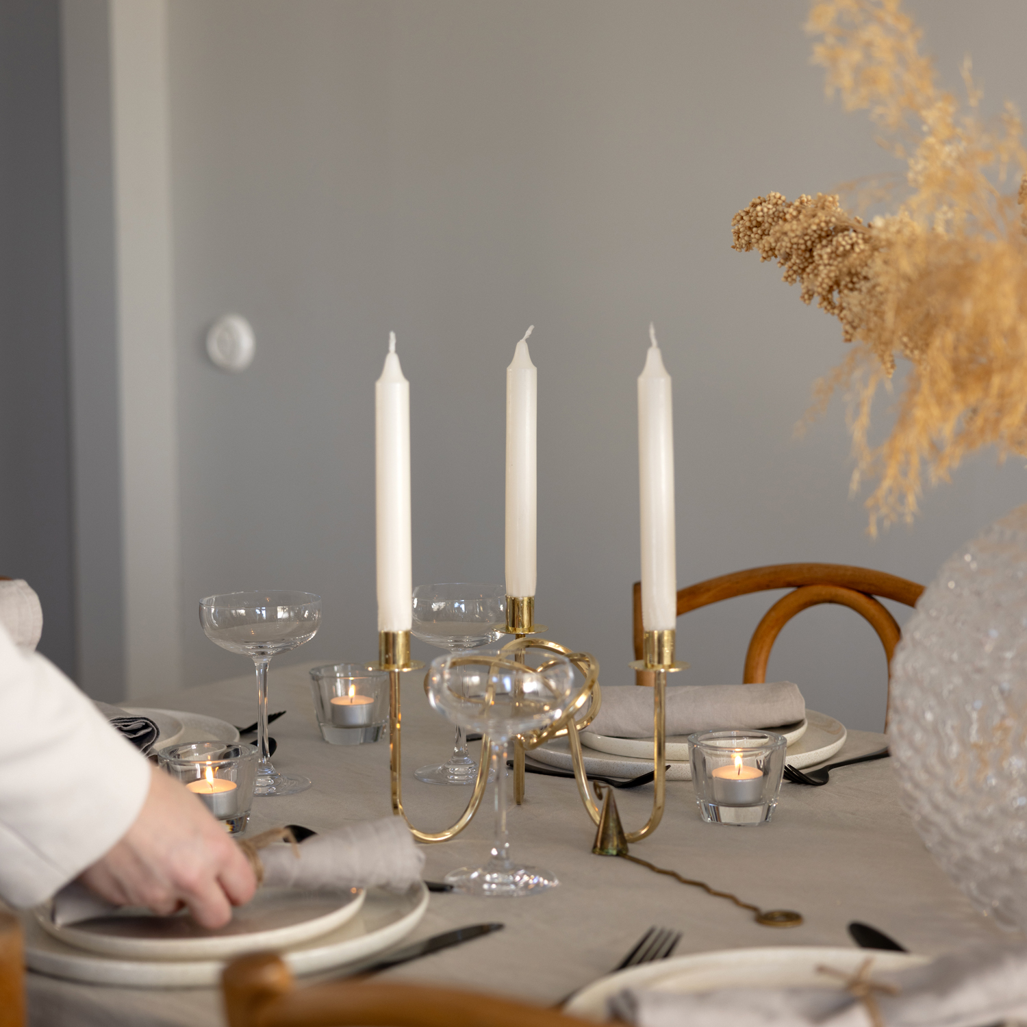 Ett elegant dukat bord med tre vita kronljus i en guldfärgad ljusstake i mitten. Ljusstakarna är ännu inte tända. På bordet står även champagneglas, vita tallrikar med grå linneservetter, värmeljus i glas och en stor vas med torkade blommor. En person i vit kavaj lägger en servett på en tallrik.