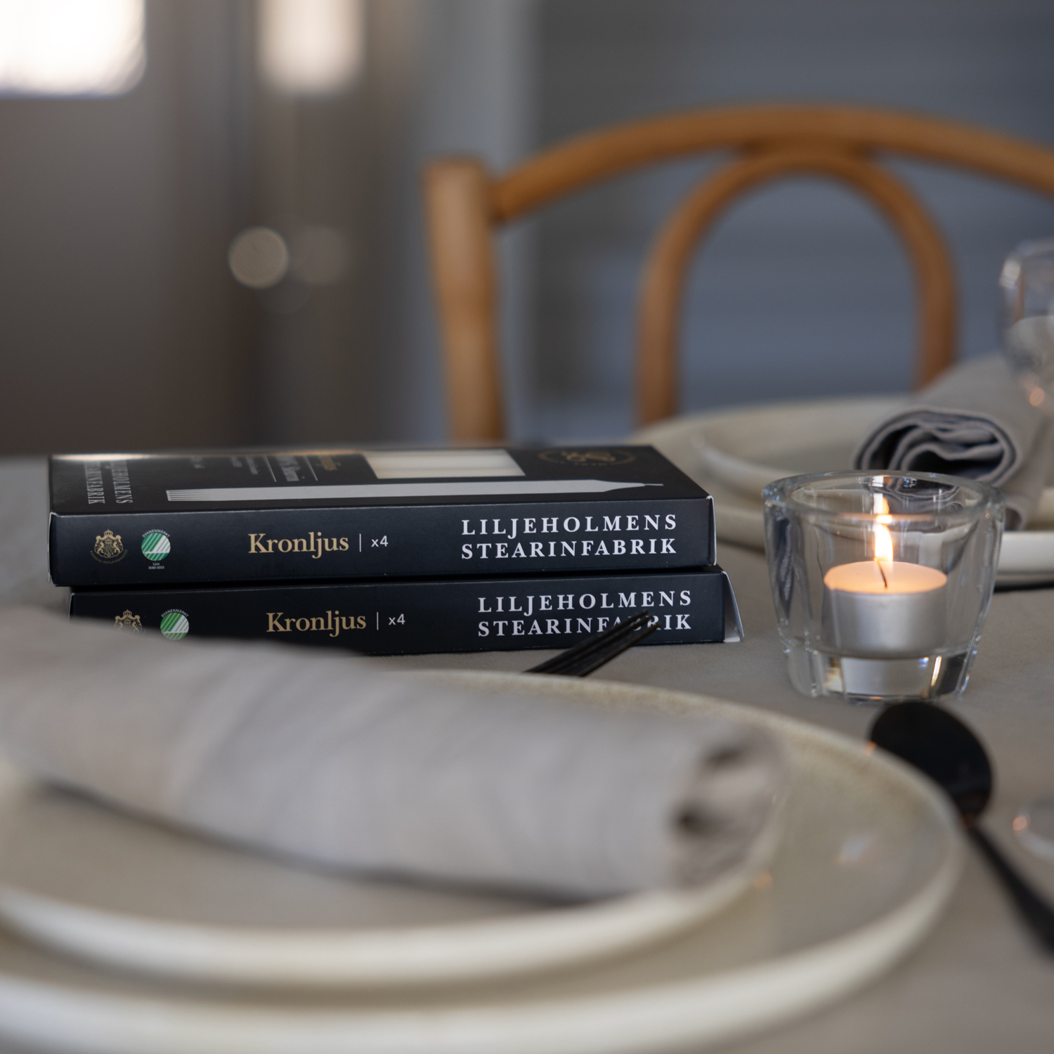 Två mörkblå förpackningar med Liljeholmens Kronljus ligger staplade på ett dukat bord. På sidan av förpackningen står ”Kronljus x4” och ”Liljeholmens Stearinfabrik” tillsammans med Svanenmärkningen. Bredvid förpackningarna står ett tänt värmeljus i ett glas. I förgrunden syns en tallrik med en grå linneservett och bestick.