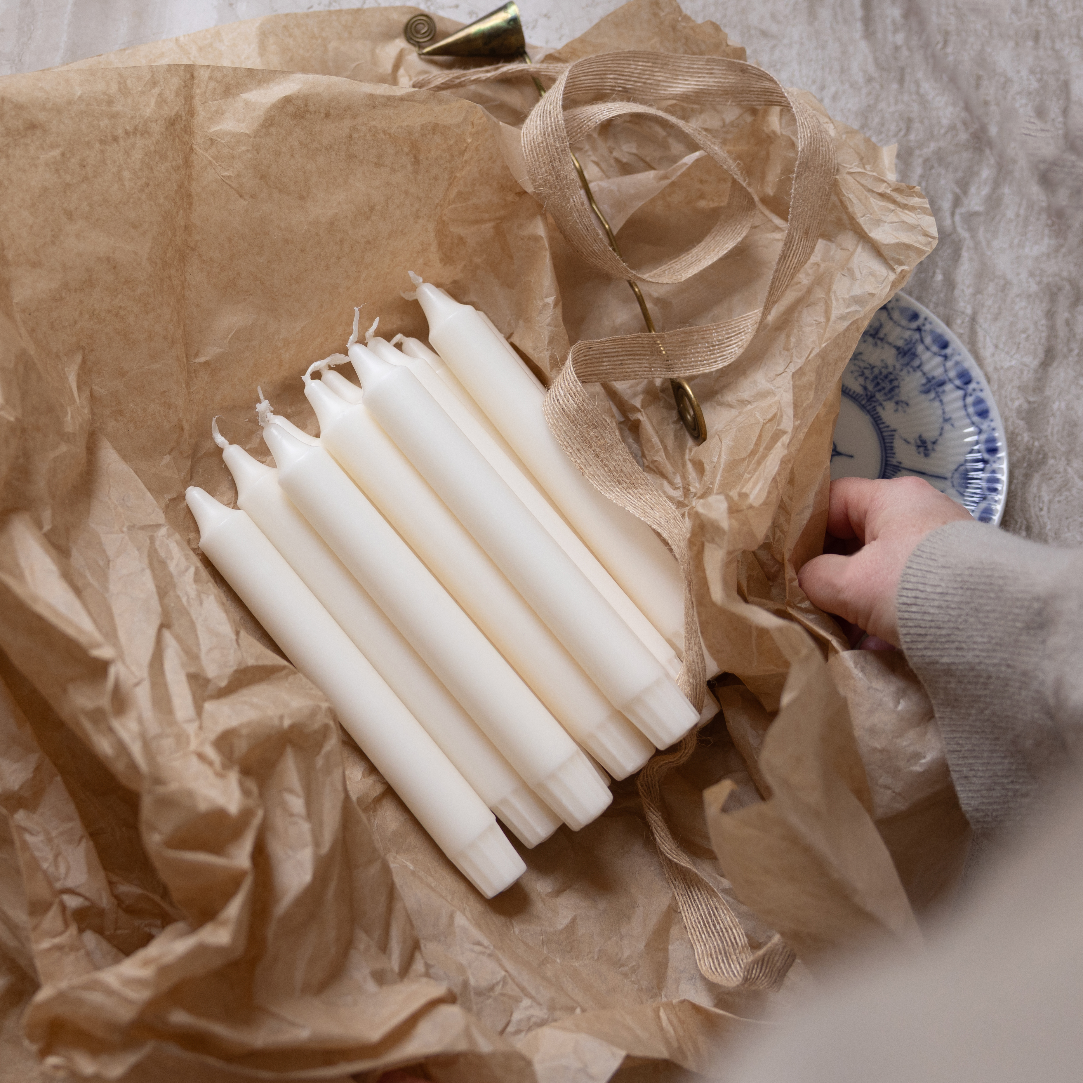Ett knippe vita kronljus ligger på brunt silkespapper. Bredvid ligger ett beige band med en ljussläckare i metall, och en hand i grå tröjärm håller i kanten av pappret. En blåvit porslinstallrik skymtar i bakgrunden.