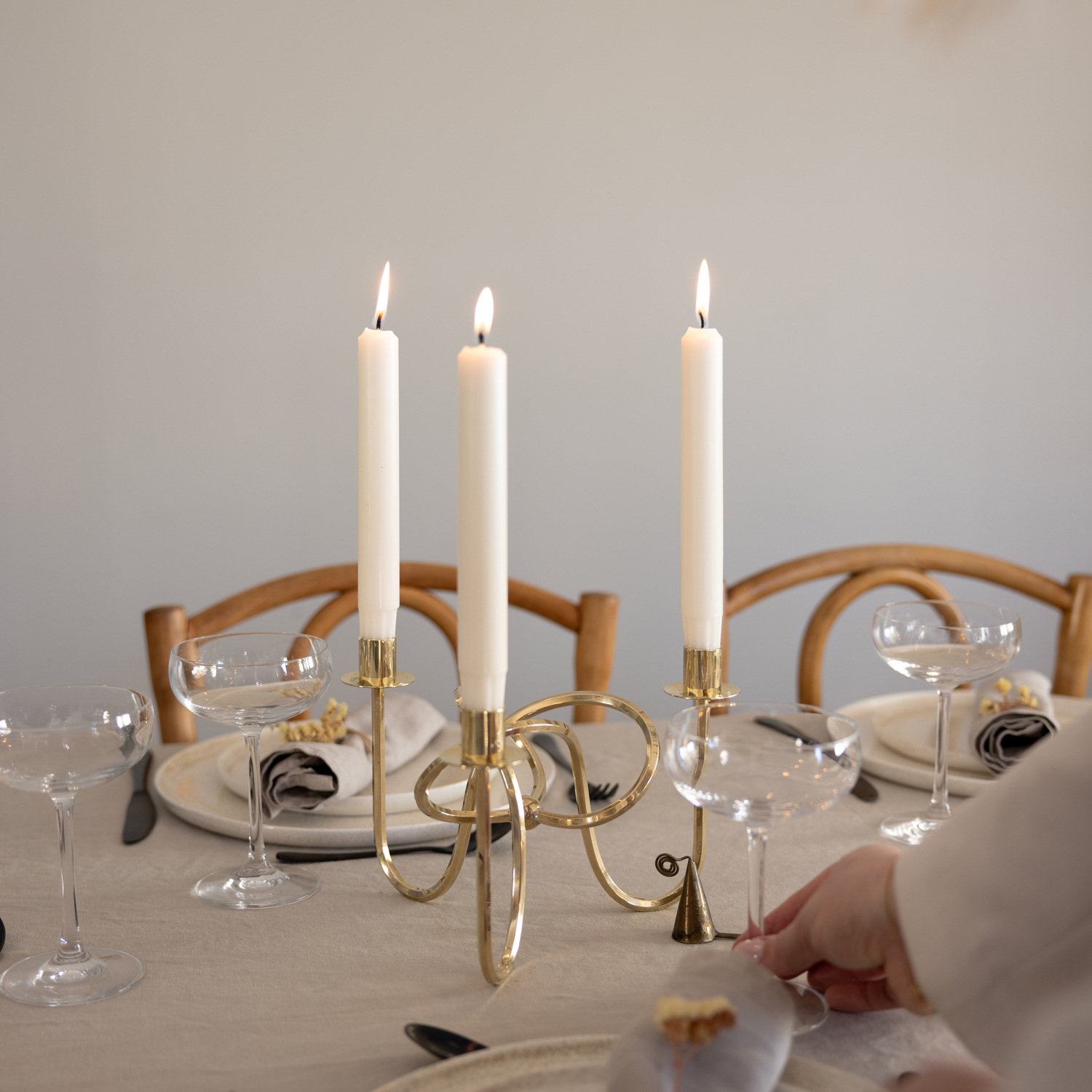 Tre vita kronljus brinner i en guldfärgad ljusstake mitt på ett dukat matbord. Bordet är täckt med en beige duk och dekorerat med tallrikar, linneservetter, champagneglas och svarta bestick. I förgrunden syns en hand som justerar ett glas.