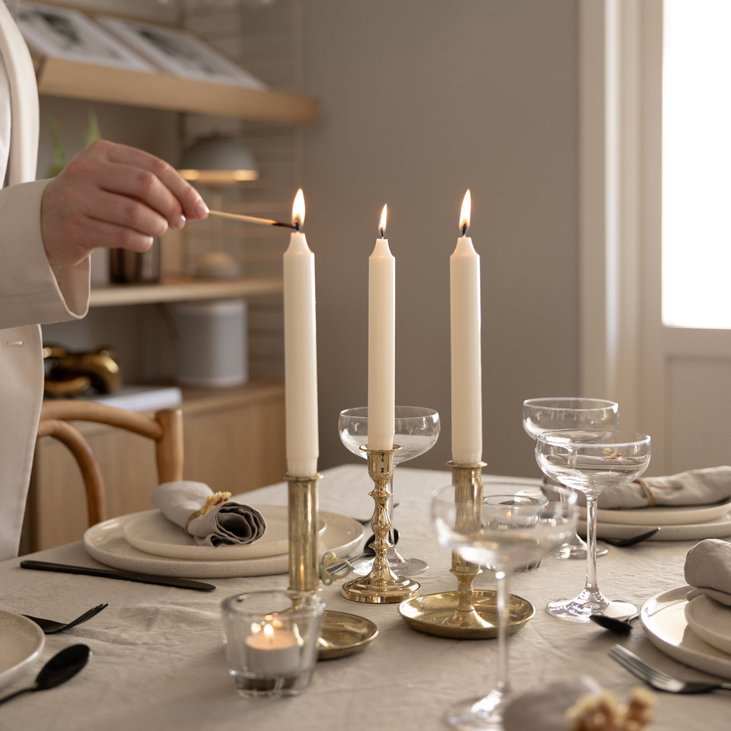Tre vita kronljus står tätt tillsammans i guldfärgade ljusstakar på ett elegant dukat bord. En person i ljus kavaj tänder det sista ljuset med en tändsticka. På bordet finns vita tallrikar, linneservetter, champagneglas och små värmeljus. I bakgrunden syns en hylla med dekorationer.