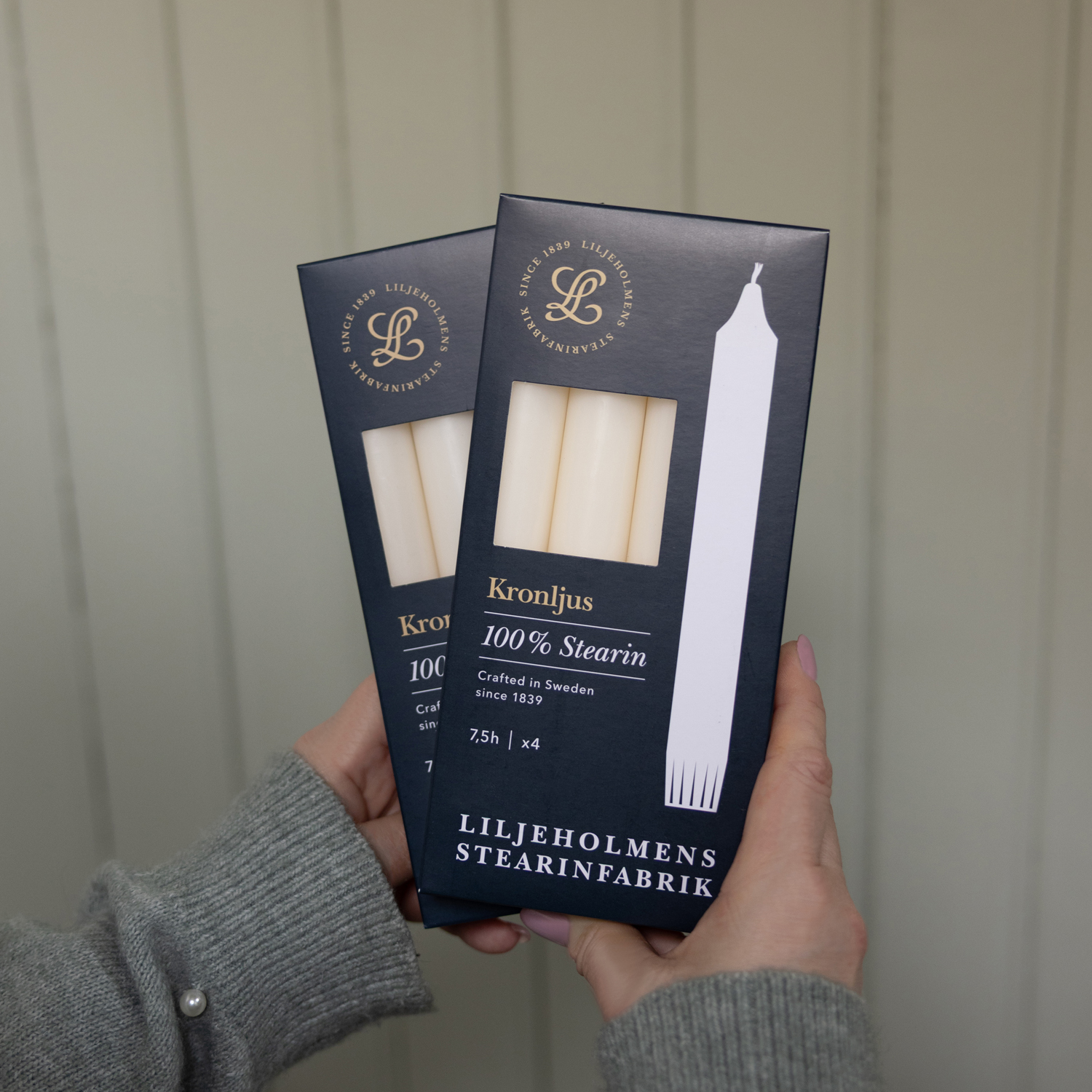 Två händer håller i två förpackningar med vita kronljus från Liljeholmens Stearinfabrik. Förpackningarna är mörkblå med guld- och vit text, där det står "Kronljus – 100 % stearin – Crafted in Sweden since 1839". Bakgrunden är ljusgrå med panelstruktur.