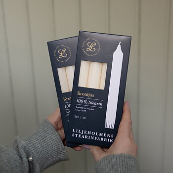 Två händer håller i två förpackningar med vita kronljus från Liljeholmens Stearinfabrik. Förpackningarna är mörkblå med guld- och vit text, där det står "Kronljus – 100 % stearin – Crafted in Sweden since 1839". Bakgrunden är ljusgrå med panelstruktur.