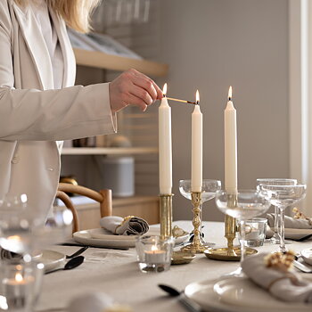 Ett dukat middagsbord med tre vita kronljus i mässingsljusstakar. En person i ljus kavaj tänder det mittersta ljuset med en tändsticka. På bordet finns också glas, tallrikar med gråa linneservetter och små värmeljus i glas.