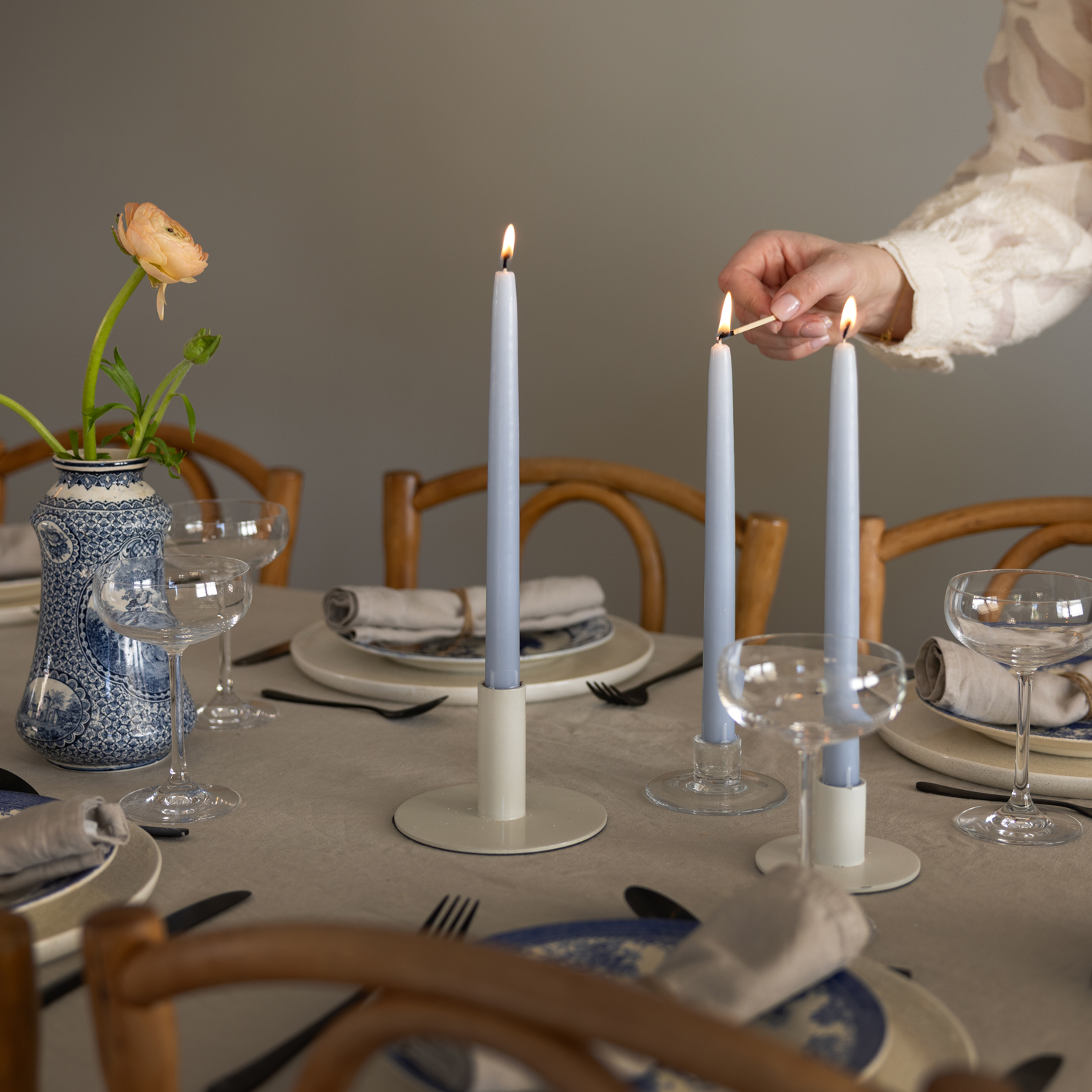 Ett elegant dukat bord med tre ljusblå antikljus som håller på att tändas av en person i ljus blus. På bordet står coupeglas, tallrikar med vikta linneservetter, svarta bestick och en blåvit vas med en aprikosfärgad ranunkel.