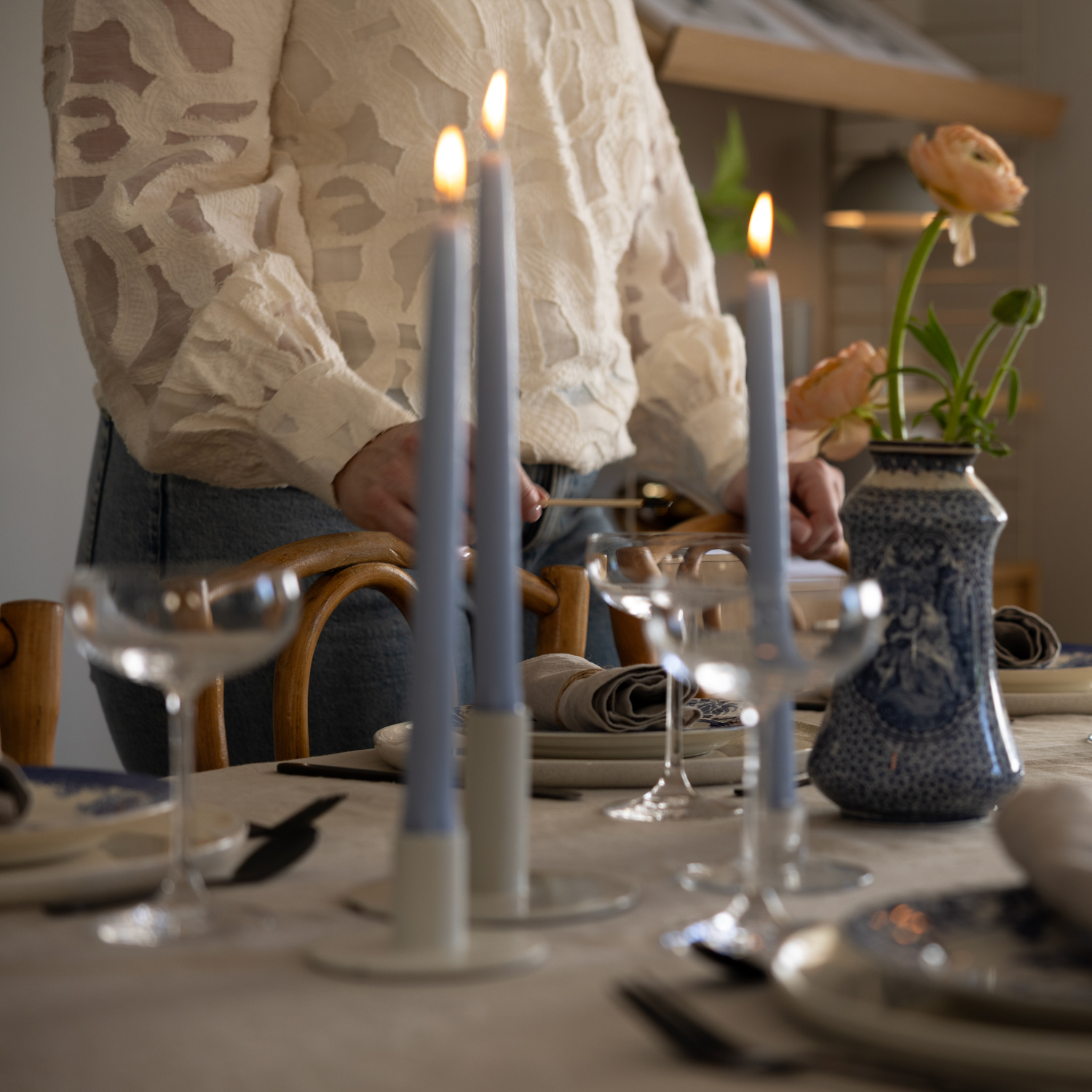 En person i ljus mönstrad blus och jeans dukar ett bord med tända ljusblå antikljus i ljusstakar. På bordet står coupeglas, tallrikar med linneservetter och en blåvit vas med aprikosfärgade ranunkler.