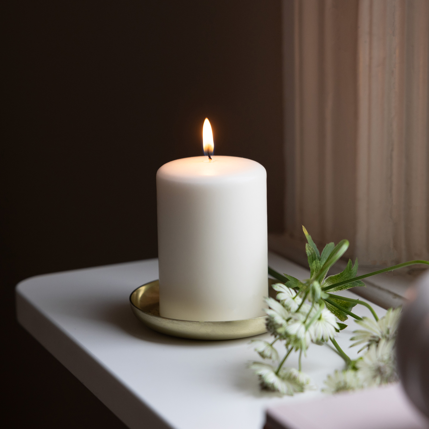 Ett tänt vitt blockljus står på ett mässingsfärgat fat på en vit fönsterbräda. Bredvid ljuset ligger vita blommor med gröna blad. Bakgrunden är mörk och skapar en lugn och stämningsfull atmosfär.