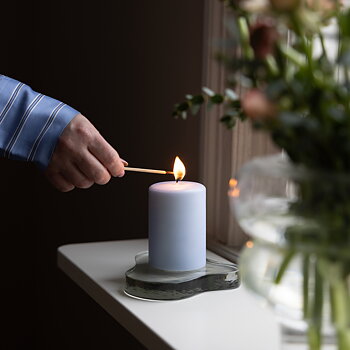 En person i blå- och vitrandig skjorta tänder ett ljusblått blockljus med en tändsticka. Ljuset står på ett glasfat med organisk form på en vit fönsterbräda. Till höger i bilden syns en suddig glasvas med snittblommor.