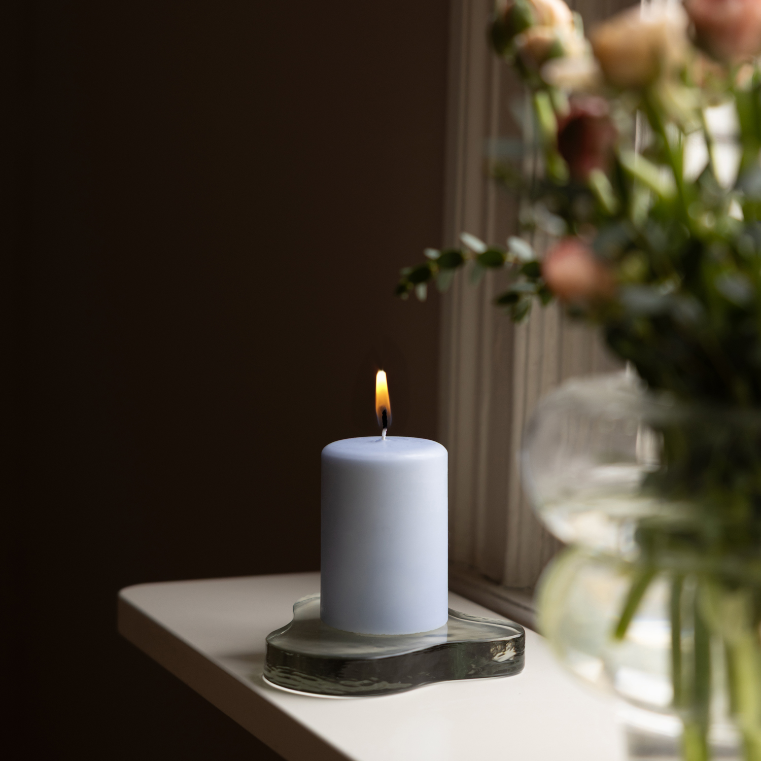 Ett tänt ljusblått blockljus står på ett organiskt format glasfat på en vit fönsterbräda. Till höger i bilden syns en suddig glasvas med snittblommor. Mörk bakgrund skapar en varm och rofylld stämning.