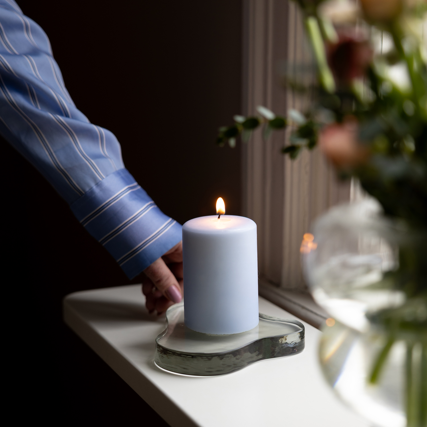 En hand i blå- och vitrandig skjorta placerar ett tänt ljusblått blockljus på ett organiskt format glasfat på en vit fönsterbräda. Till höger syns en suddig vas med snittblommor. Bakgrunden är mörk och stämningsfull.