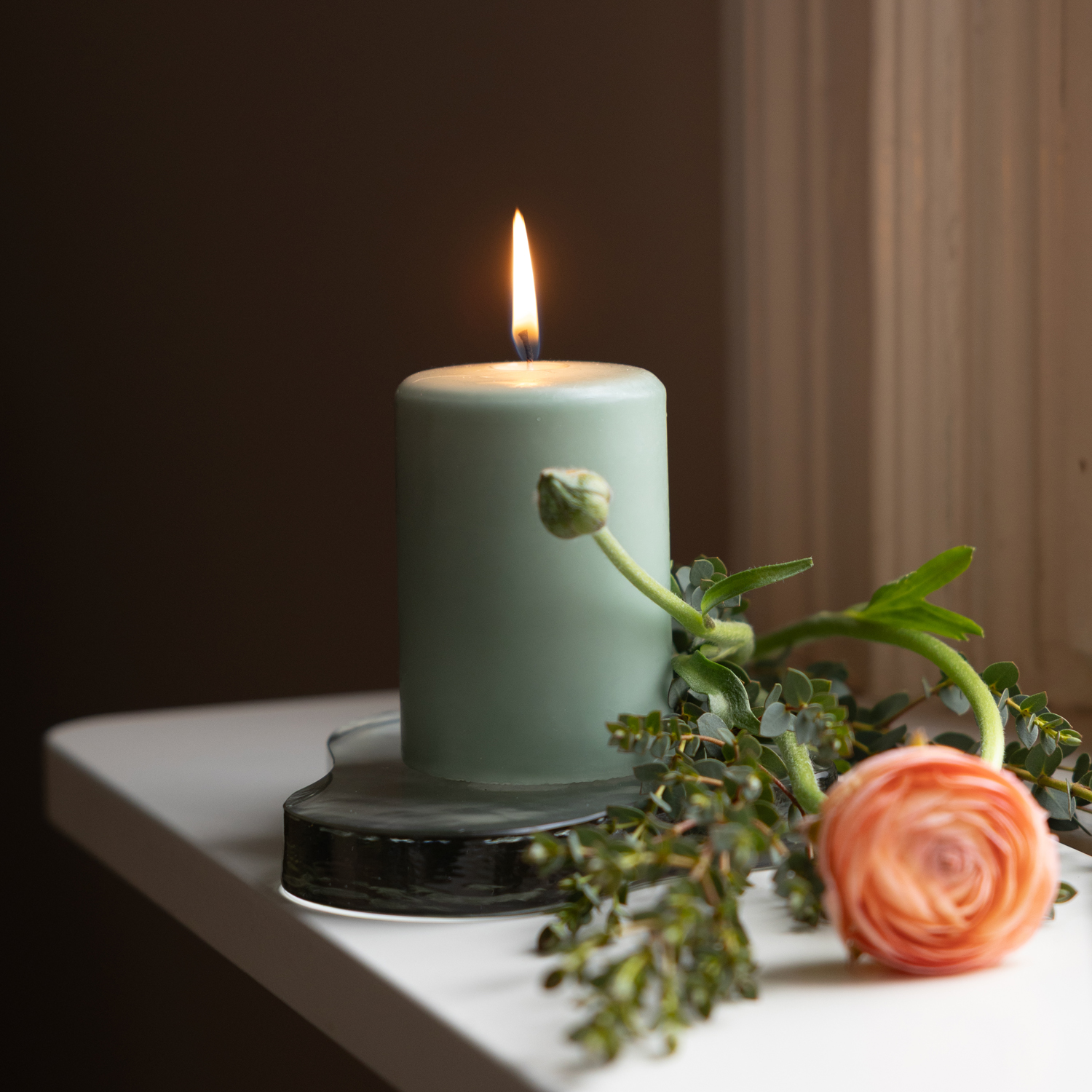 Ett tänt ljusgrönt blockljus står i ett hjärtformat ljusfat av glas på en vit fönsterbräda. Bredvid ljuset ligger gröna kvistar och en aprikosfärgad ranunkelblomma. Ljuset skapar en varm och rofylld stämning vid fönstret.