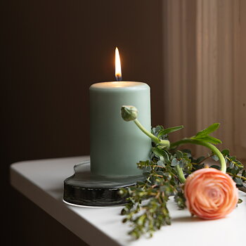 Ett tänt ljusgrönt blockljus står i ett hjärtformat ljusfat av glas på en vit fönsterbräda. Bredvid ljuset ligger gröna kvistar och en aprikosfärgad ranunkelblomma. Ljuset skapar en varm och rofylld stämning vid fönstret.