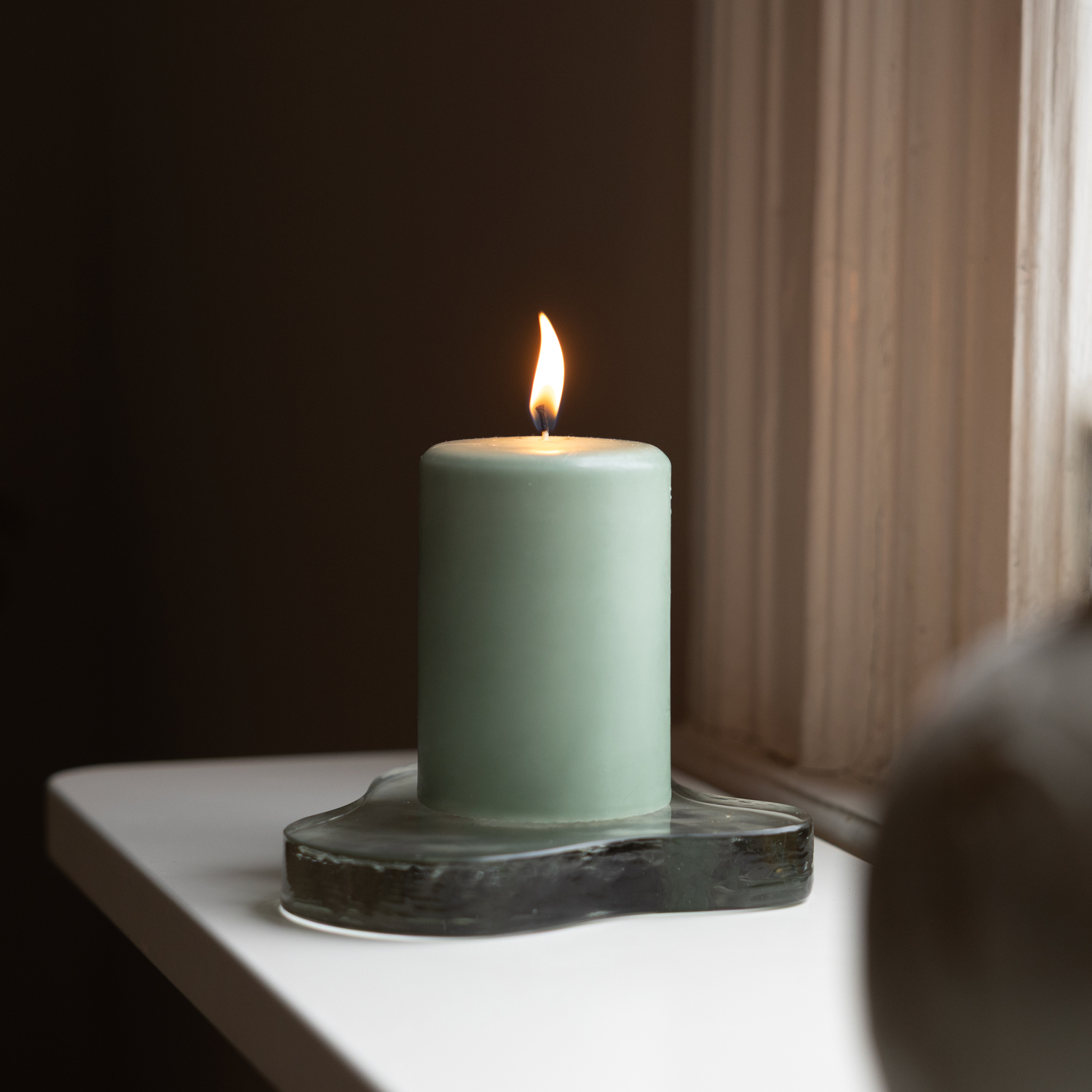 Ett tänt ljusgrönt blockljus står på ett transparent hjärtformat ljusfat i glas på en vit fönsterbräda vid ett fönster. Lågan lyser stilla och skapar en varm och lugn atmosfär mot den mörka bakgrunden.