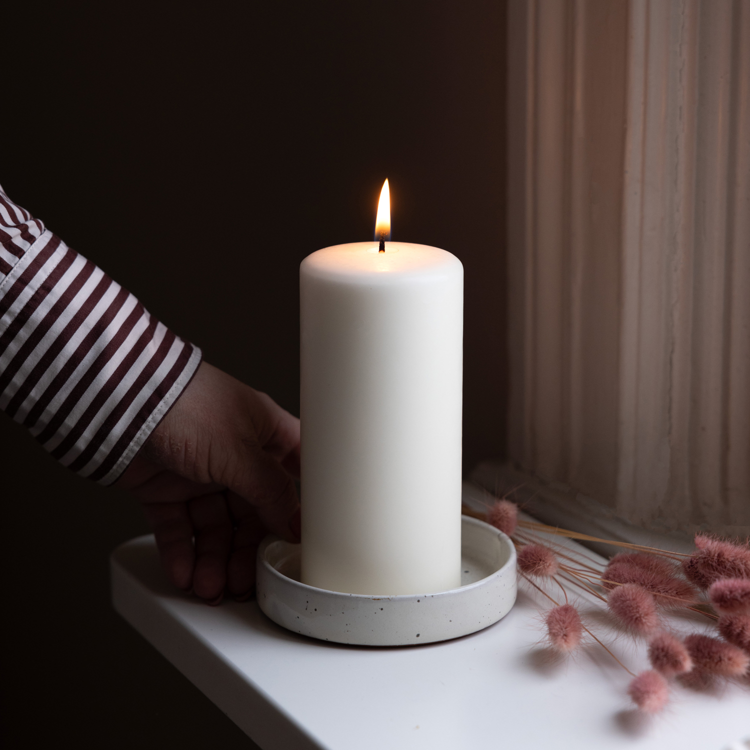 En hand i röd- och vitrandig skjorta placerar ett tänt vitt blockljus i en ljusgrå ljushållare på ett vitt fönsterbräde. Bredvid ljuset ligger rosa torkade blommor, och fönsterkarmen syns i bakgrunden. Bilden förmedlar en varm och ombonad stämning.