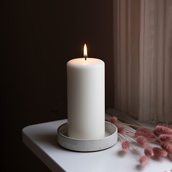 Ett tänt vitt blockljus står i en ljusgrå ljushållare på ett vitt fönsterbräde. Bredvid ljuset ligger rosa torkade blommor, och bakgrunden är mörk med ett gammalt fönster med vita karmar. Stämningen är varm och harmonisk.
