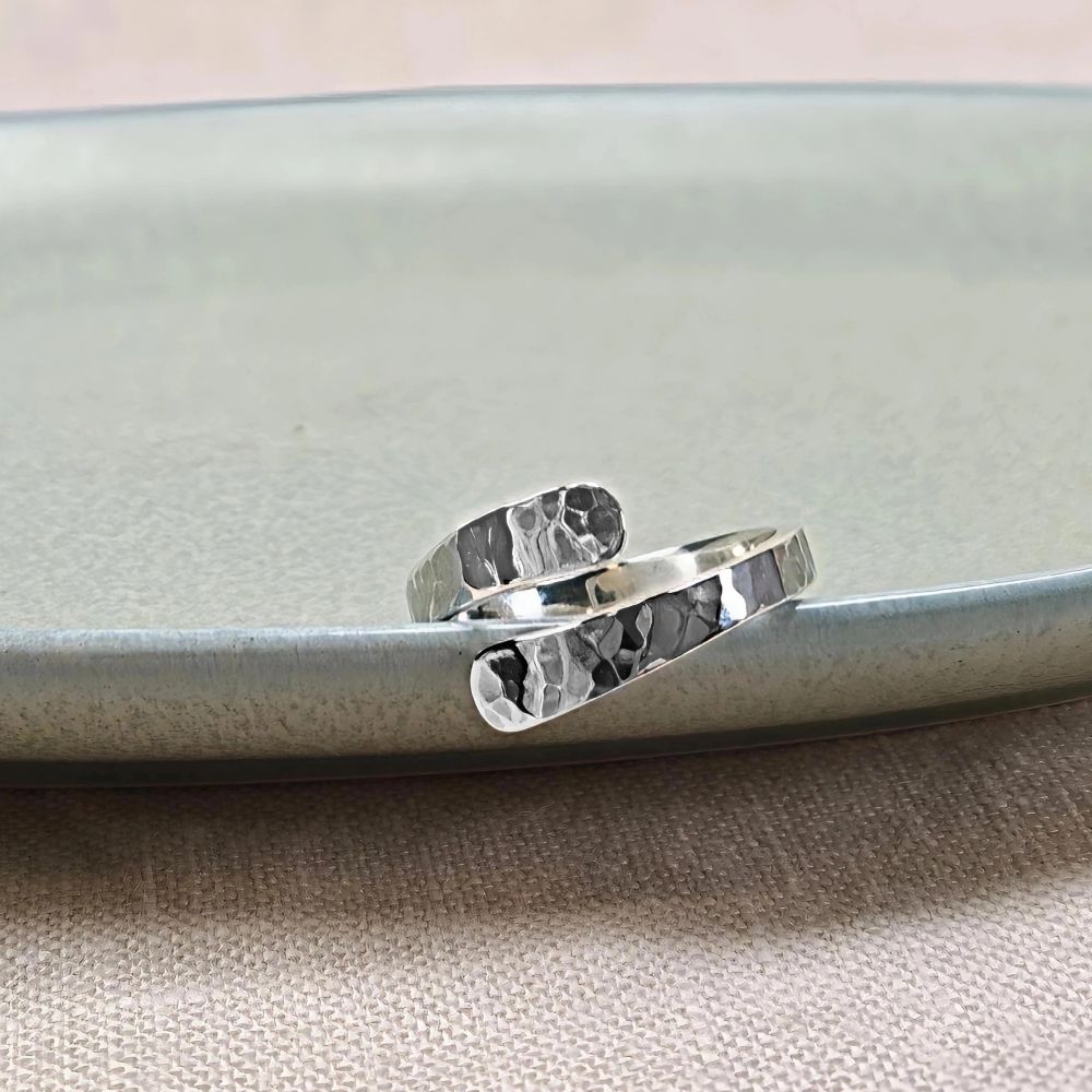 IAMLVåga ring i silver, justerbar hamrad ring med omlott design, visad liggande på ett ljusgrönt fat mot beige tygbakgrund.UND Våga ring silver