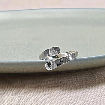IAMLVåga ring i silver, justerbar hamrad ring med omlott design, visad liggande på ett ljusgrönt fat mot beige tygbakgrund.UND Våga ring silver
