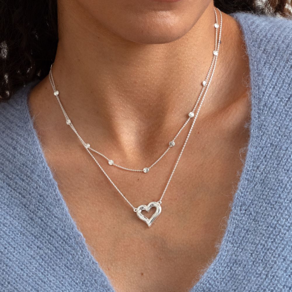 Syster P Solea halsband i silver, tunt kedjehalsband med små hamrade länkar i två lager, stylat med ett hjärthalsband i silver mot en ljusblå stickad tröja.