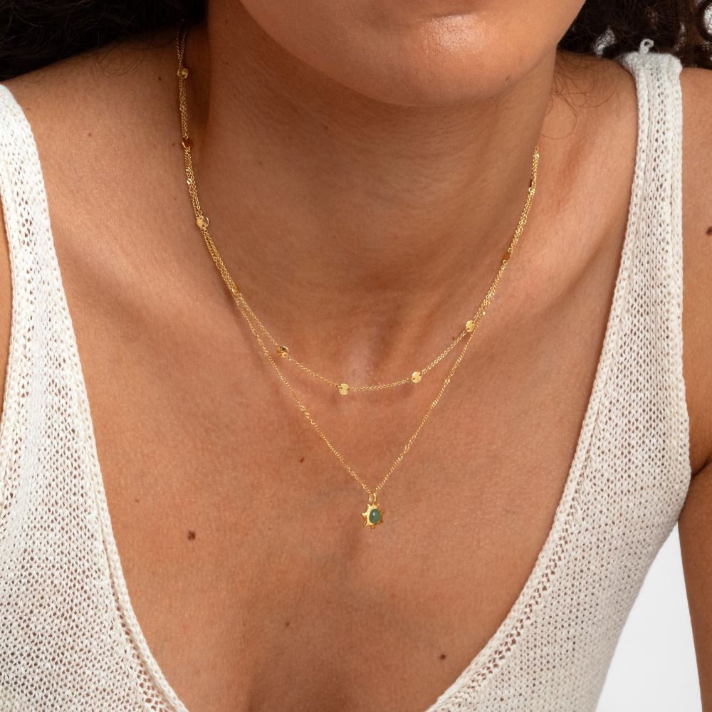 Syster P Solea halsband guld, tunt kedjehalsband med små hamrade länkar, stylat i lager med ett halsband med grön sten mot vit stickad topp.