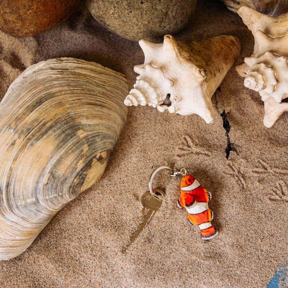 Porte-clés Poisson-clown posé sur du sable, près de coquillages et de pierres.
