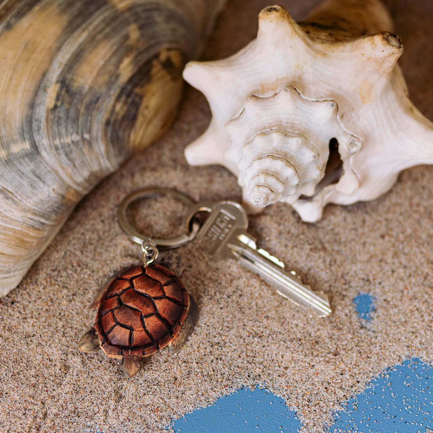 Porte-clés Tortue de mer posé sur du sable avec des coquillages en arrière-plan.