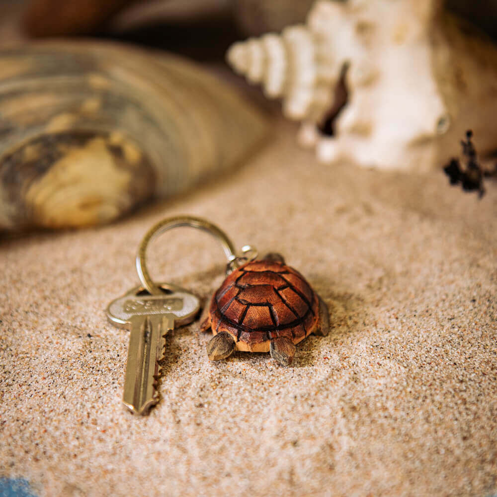 Porte-clés Tortue de mer posé sur du sable à côté de coquillages.