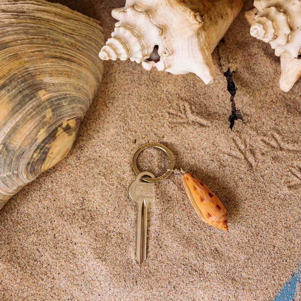 Porte-clés Pétoncle posé sur du sable à côté de coquillages.