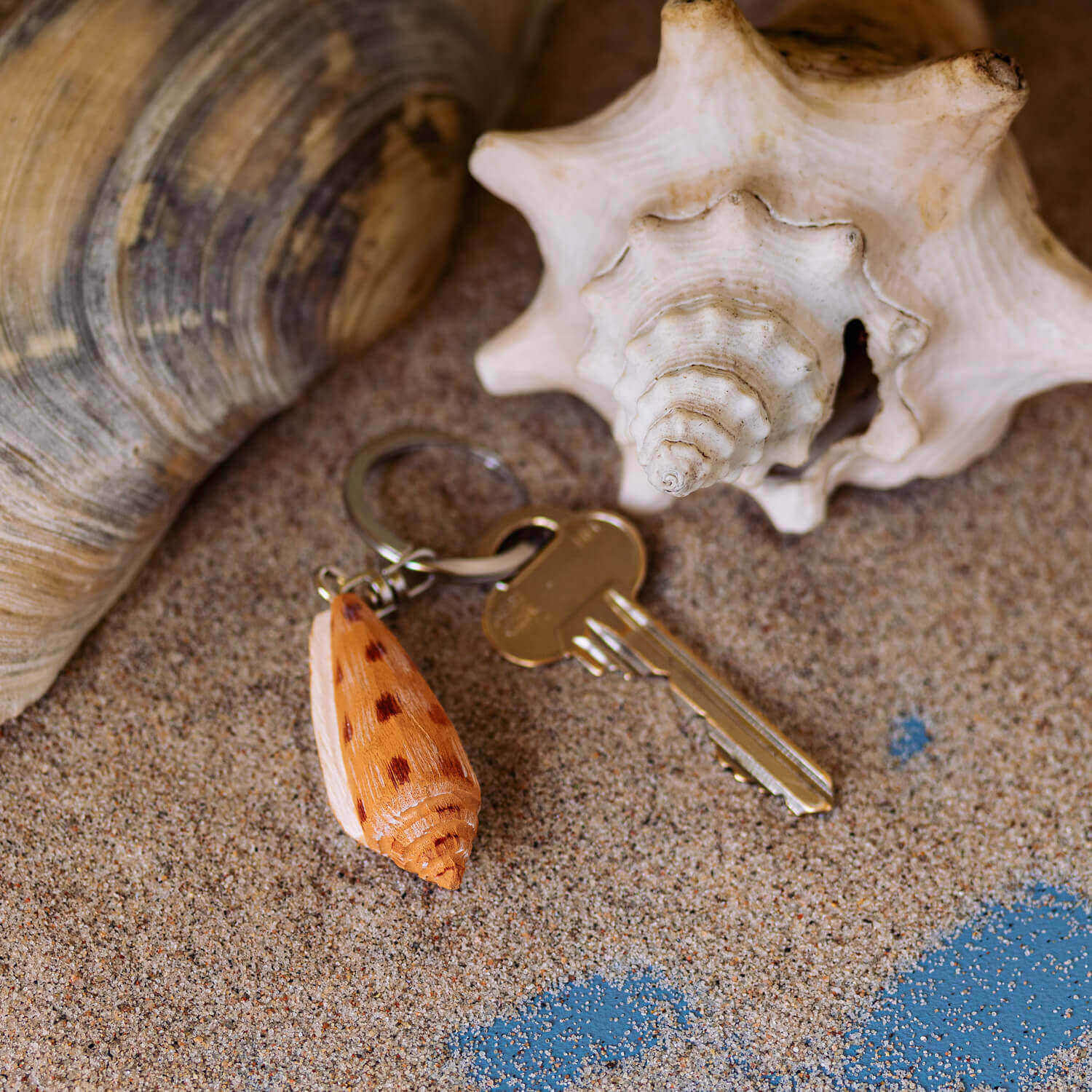 Porte-clés Pétoncle posé sur du sable avec des coquillages en arrière-plan.