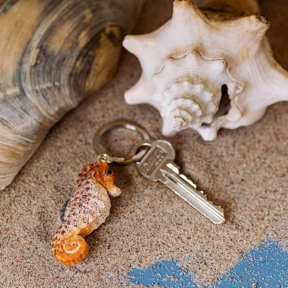 Porte-clés Hippocampe avec une hippocampe sculptée à la main posée sur du sable parmi des coquillages.